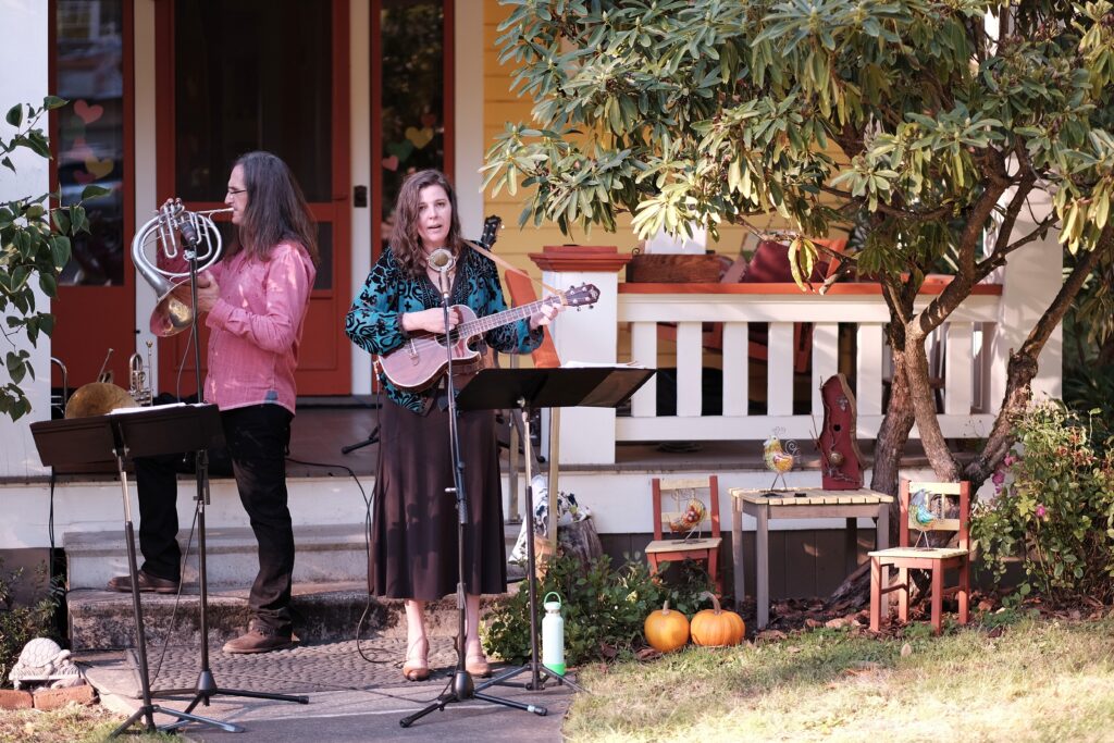 man on horn and woman on guitar play instruments in front of a porch